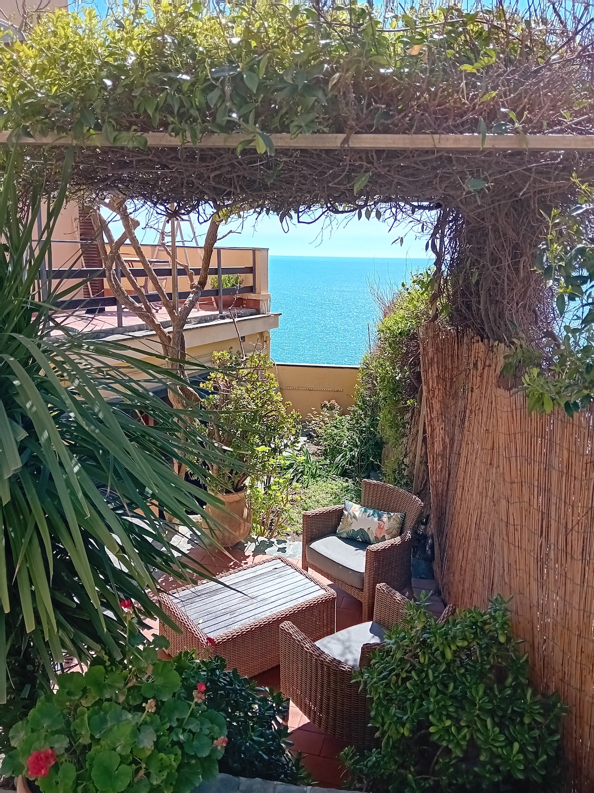 A private outdoor patio is surrounded by lush greenery and features comfortable seating. A low coffee table sits between two chairs, with a backdrop of the serene Mediterranean Sea visible in the distance, enhancing the relaxed atmosphere of the space.