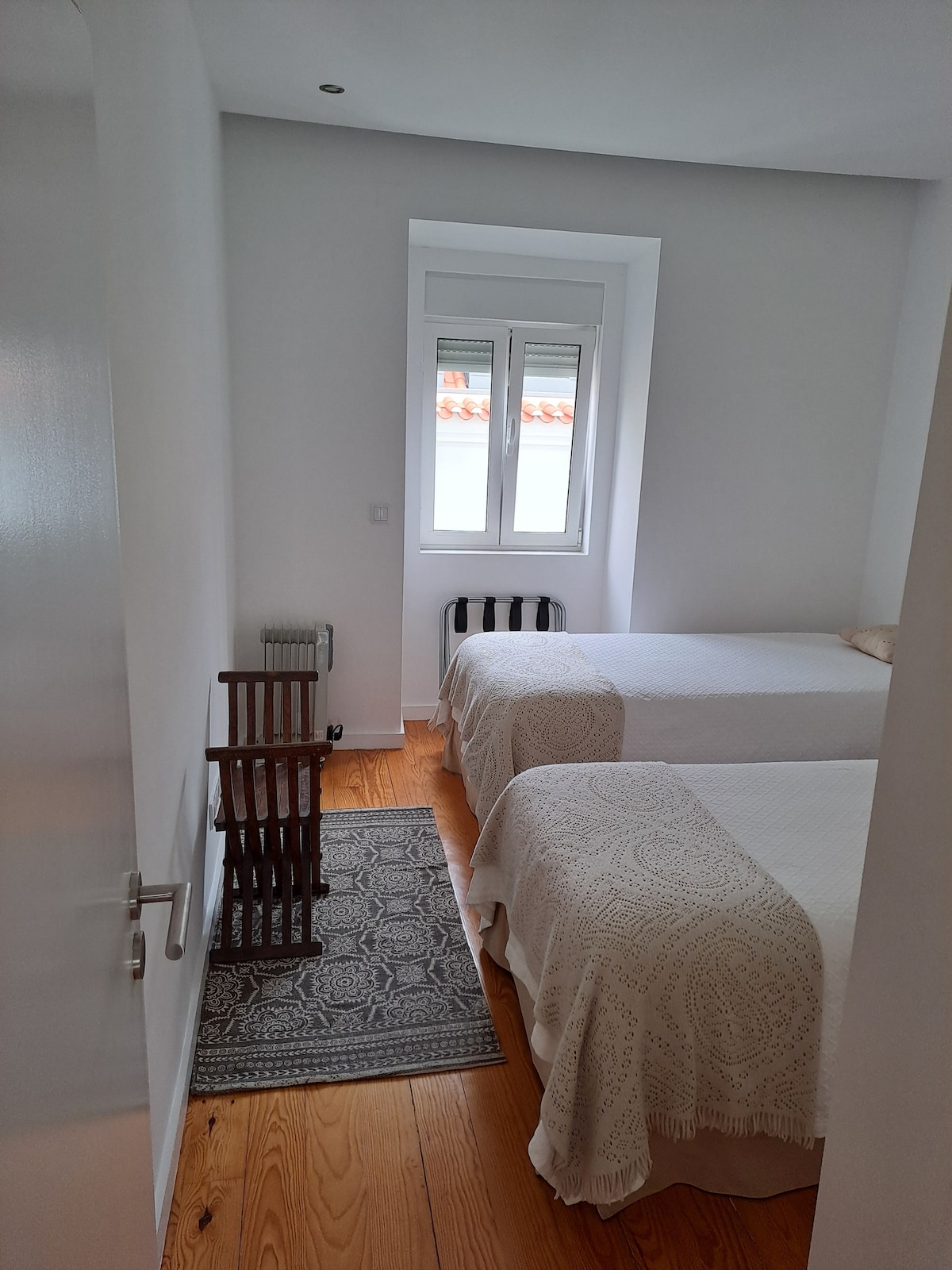 The bedroom features two single beds with light-colored bedding and decorative throws. A woven rug is positioned between the beds, adding texture. Natural light enters through a small window with white frames, illuminating the light-colored walls and wooden floor.