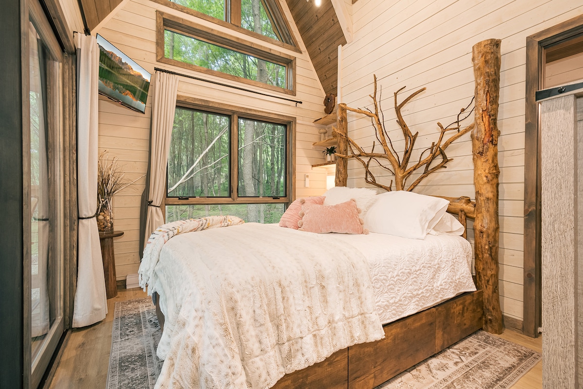 A serene bedroom features a spacious bed adorned with white linens and decorative pillows. Large windows offer abundant natural light and views of the forest outside. Wooden accents, including a tree trunk headboard, contribute to the rustic aesthetic, complemented by a patterned area rug.