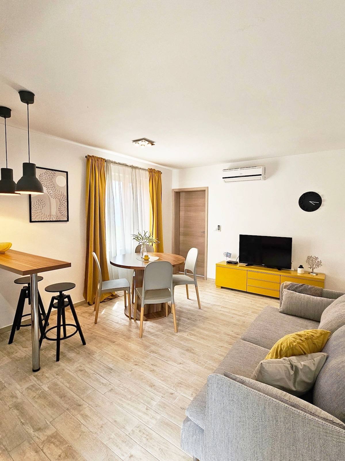 The living area features a cozy gray sofa with decorative pillows, alongside a wooden dining table surrounded by four light-colored chairs. A flat-screen television sits on a bright yellow console, while warm curtains diffuse natural light from the nearby window. Pendant lights hang above the bar area.