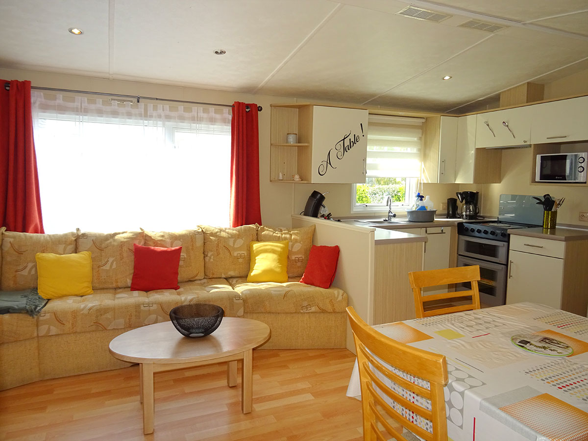 A cozy living area features a beige sofa adorned with colorful cushions. Natural light enters through a large window, complemented by light curtains. A dining table with wooden chairs is set in the foreground, while an open kitchen equipped with modern appliances is visible in the background.