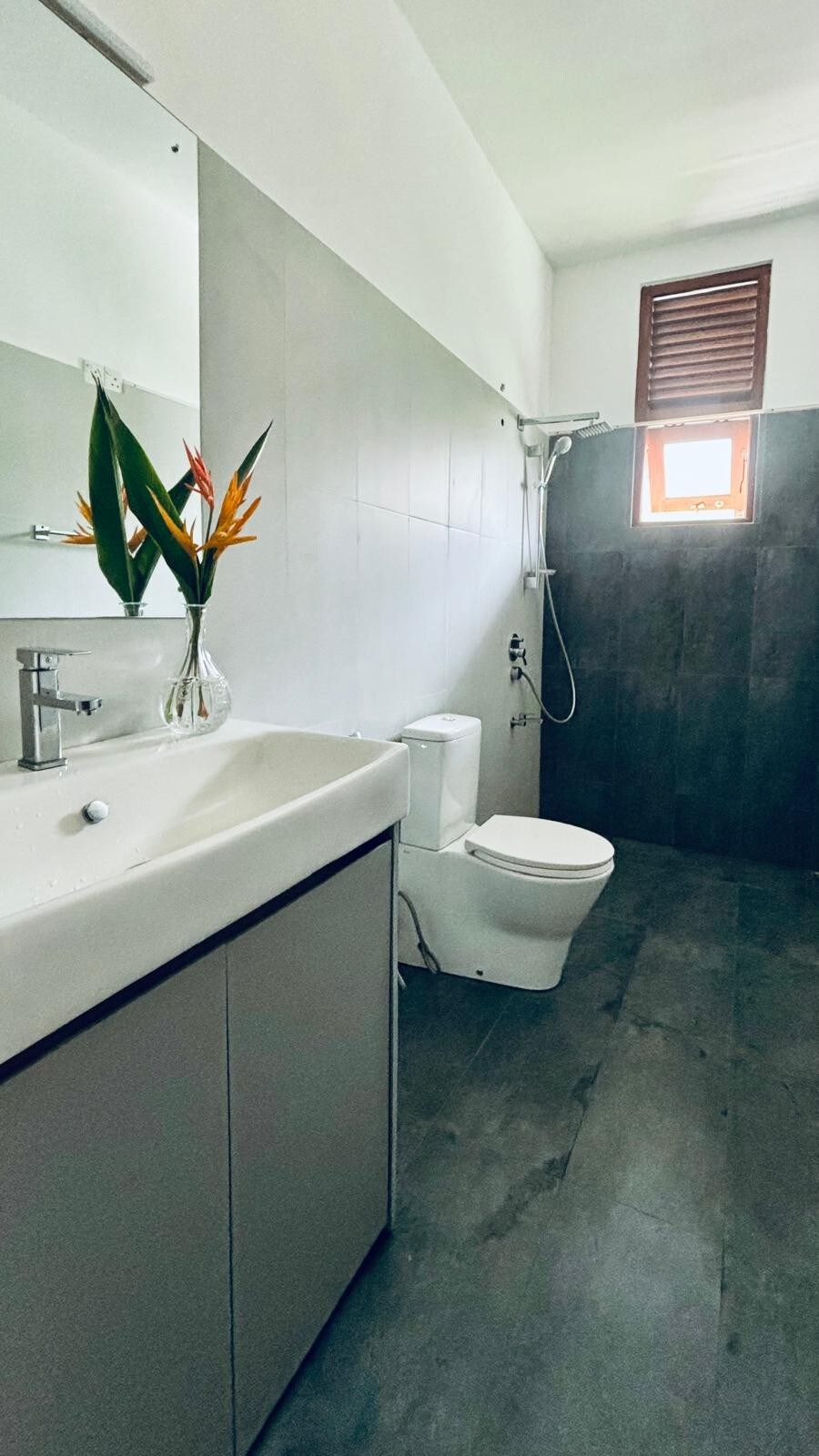 A modern bathroom features a sleek vanity with a single sink and ample storage beneath. A shower is set against a tiled wall, while a small window allows natural light to enter. A vase with tropical flowers adds a touch of color to the space.