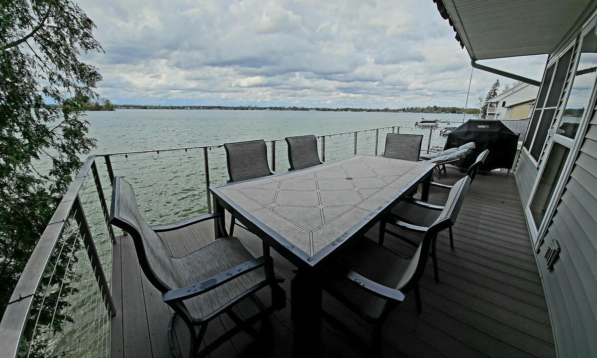 An outdoor deck offers a large rectangular table surrounded by six chairs, ideal for dining. The railing provides unobstructed views of the tranquil lake, while a barbecue grill is positioned nearby for outdoor cooking. The sky is partly cloudy, reflecting on the water.