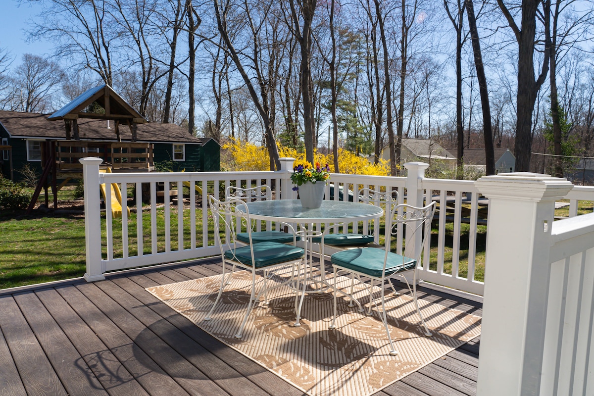 A small outdoor dining area is featured, consisting of a round glass table surrounded by four metal chairs with teal cushions. A potted plant with blooms sits at the center of the table. The deck overlooks a spacious yard, with trees in the background.