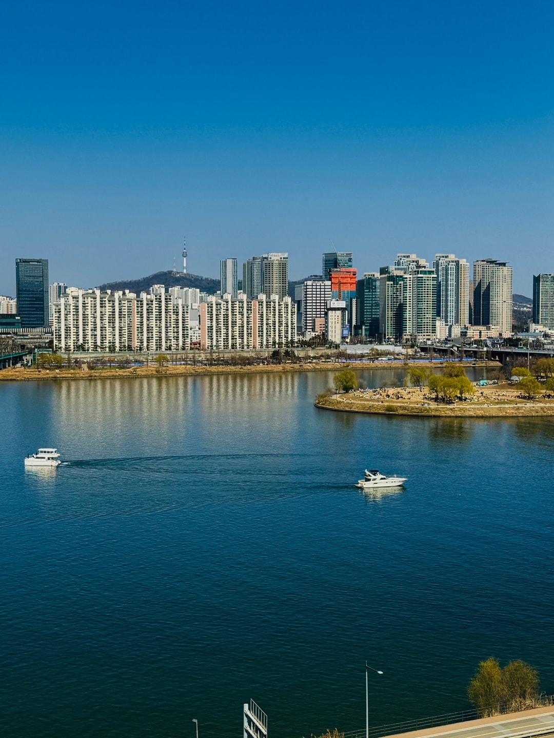 A scenic view of the Han River is captured, with a bustling city skyline visible in the background. Two boats navigate the calm waters, reflecting the surrounding buildings and a clear blue sky. The vibrant urban landscape is complemented by gently sloping hills.