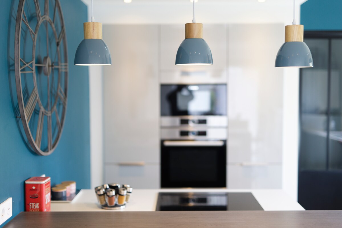 A modern kitchen is highlighted by three pendant lights with blue shades and wooden accents. A sleek oven and stove are elegantly integrated into the cabinetry, while a decorative wall clock adds interest. Items are neatly arranged on the countertop, creating an organized workspace.