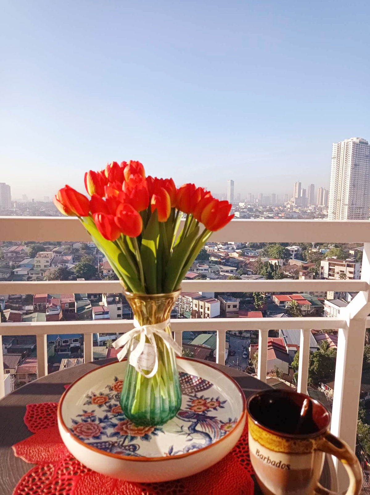 A vase filled with red tulips is positioned on a table. A printed plate rests next to a mug, with a view of the city skyline visible through the balcony railing. The scene captures a serene morning atmosphere with clear skies.