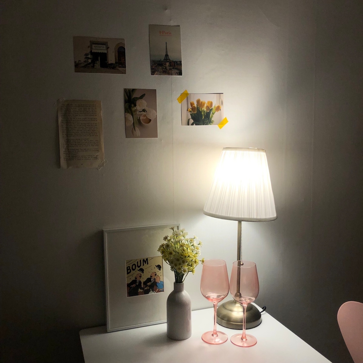A well-lit corner table displays a stylish lamp with a white pleated shade, casting a warm glow. Pink glasses and a simple vase with fresh flowers add a touch of elegance. Art prints adorn the wall above, contributing to a cozy ambiance.