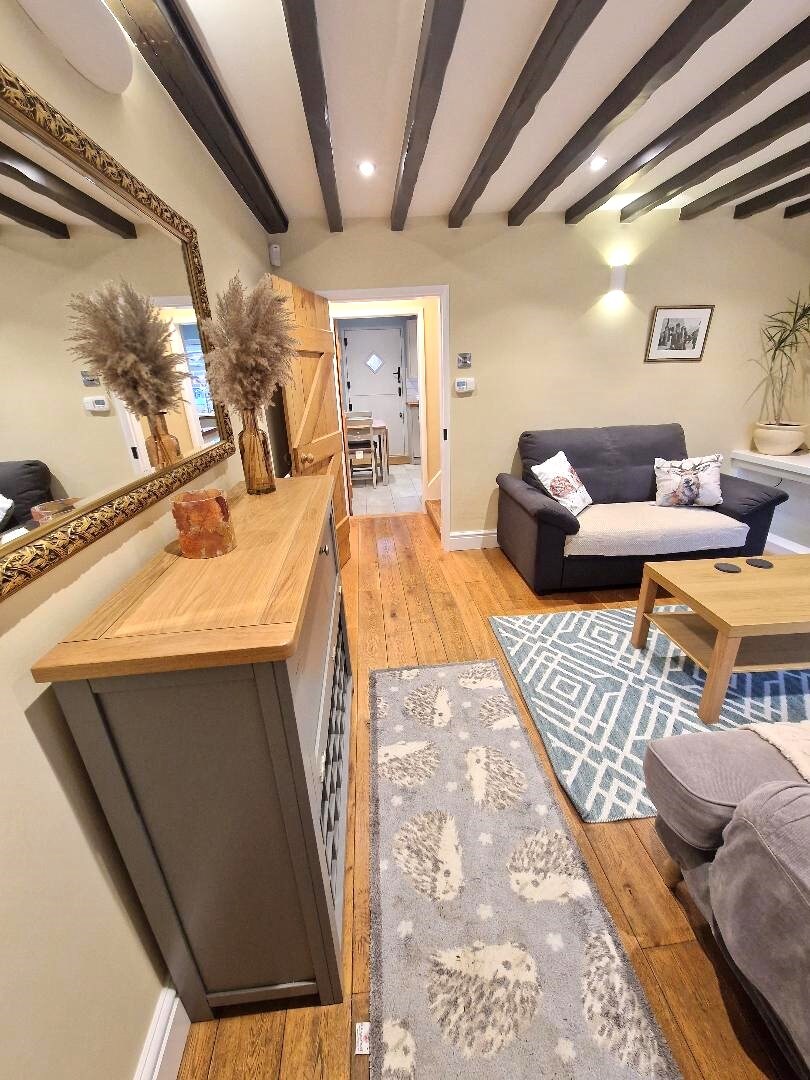 A welcoming lounge area features wooden flooring and exposed beams. A stylish console table displays decorative elements. A gray sofa is positioned opposite a low coffee table, with a textured rug underfoot. Soft lighting enhances the inviting atmosphere, while doorways lead to other spaces.