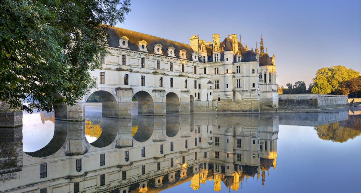 A historic château is reflected in calm water, showcasing its architectural symmetry and detailing. Lush greenery is observed along the edges, enhancing the serene environment. The building's ornate towers and windows are highlighted in the early morning light, creating a peaceful scene.