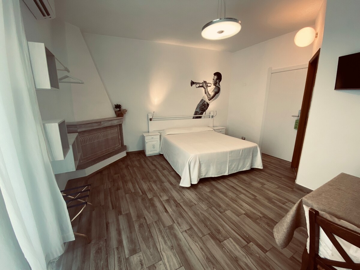 A spacious room featuring a large bed with a light cover, complemented by a decorative wall mural of a trumpet player. Natural light floods the space through a large window, while wooden flooring adds warmth. A small dining table and a chair are positioned in the corner.