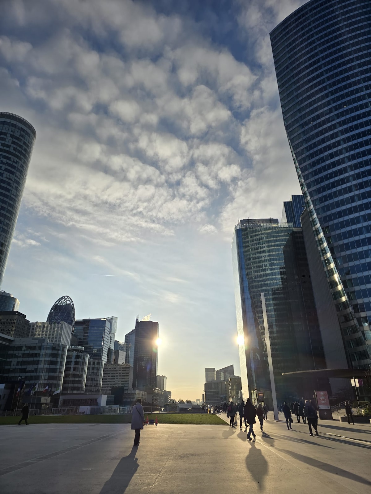 A cityscape is depicted, featuring modern skyscrapers reflecting sunlight under a partly cloudy sky. The ground level showcases people walking in the foreground, contributing to a vibrant urban atmosphere. The tall structures create a dramatic silhouette against the horizon.