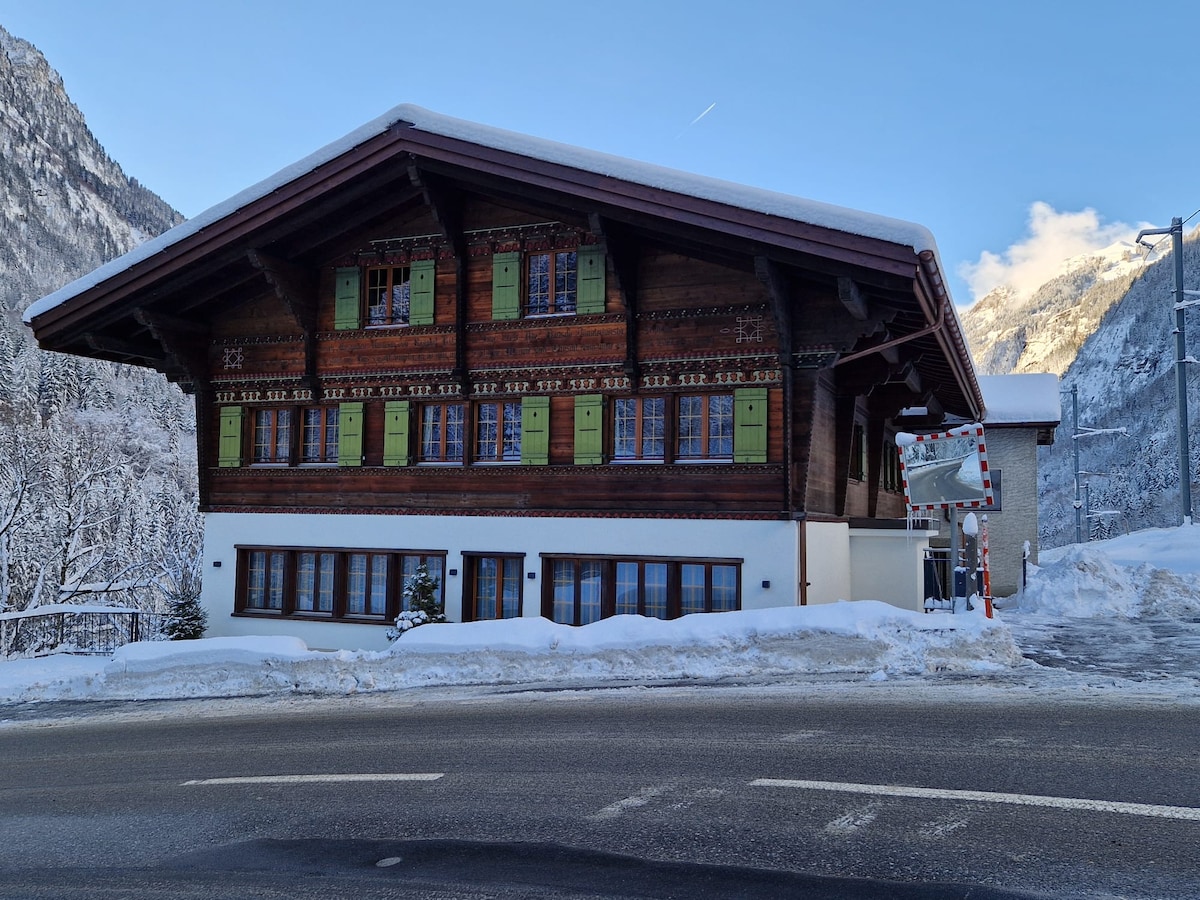 A traditional chalet features a wooden exterior adorned with green shutters and intricate detailing. Snow blankets the ground surrounding the building, while adjacent mountains are visible in the background. The structure stands alongside a paved road, highlighting its accessibility.
