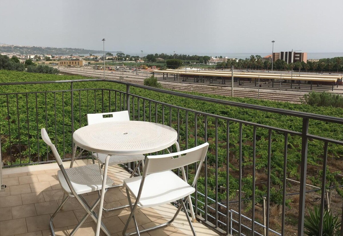 A private terrace is visible, featuring a round table paired with two white chairs. The area offers views of lush greenery and distant coastal scenery, with a glimpse of a train station in the background. The space is designed for leisurely outdoor dining.