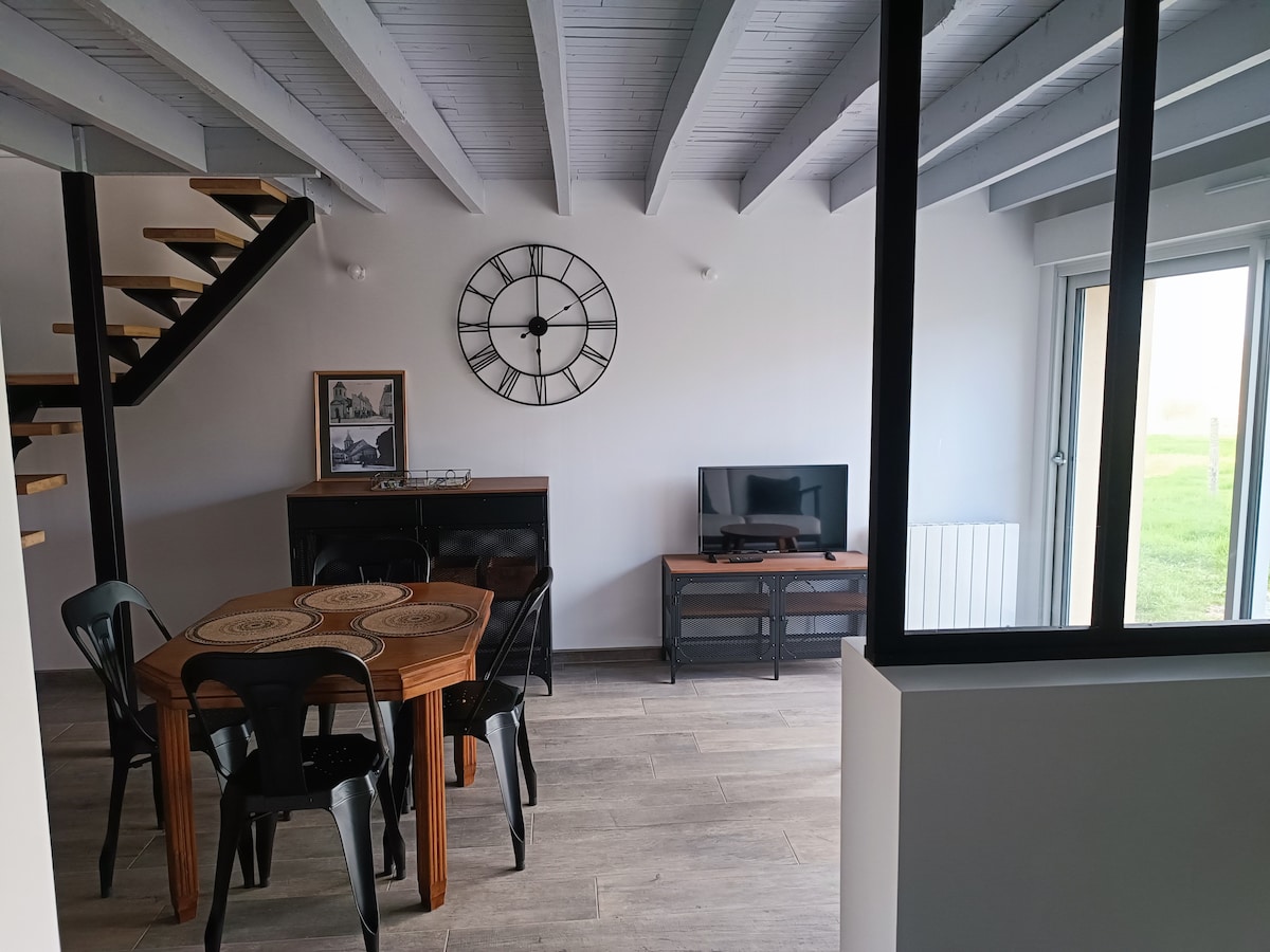 An inviting living space features a circular dining table surrounded by black chairs. A wall clock is mounted above the table, and a television is placed on a low media unit. Large glass doors lead to a view of the outdoors, enhancing the room's openness.