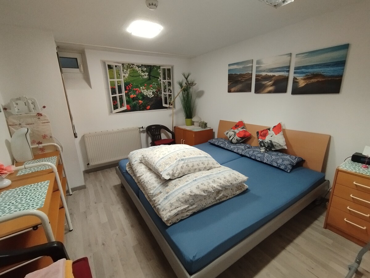The sleeping area features a spacious bed with layered bedding and decorative pillows. Above the bed, three artworks depicting serene beach scenes are displayed. Natural light enters through a window adorned with floral curtains, and a wooden dresser and chair complete the arrangement.