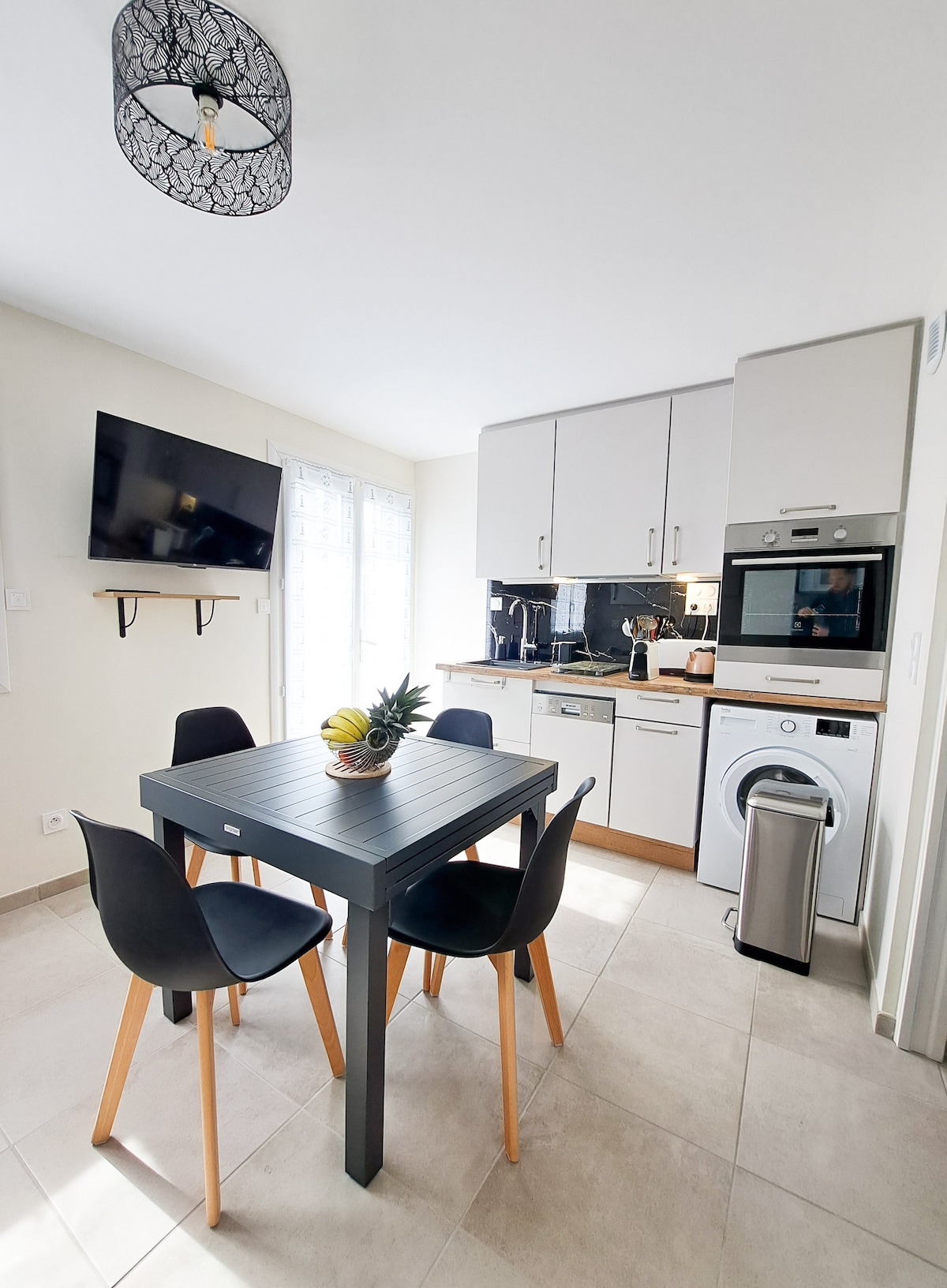 A modern kitchen area features a sleek table with four black chairs and a fruit centerpiece. An integrated stove and oven are complemented by a microwave and dishwashing unit. A wall-mounted television is visible, along with a window allowing natural light to brighten the space.