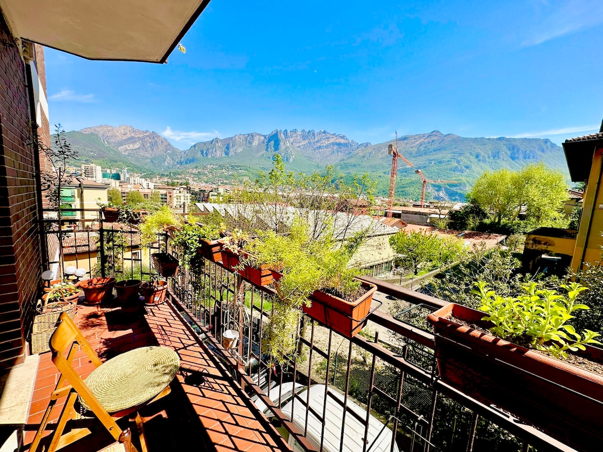 A balcony is equipped with several potted plants, providing a natural touch. Views of surrounding mountains and the city are visible in the background, with clear blue skies and sunlight casting a warm glow over the space.