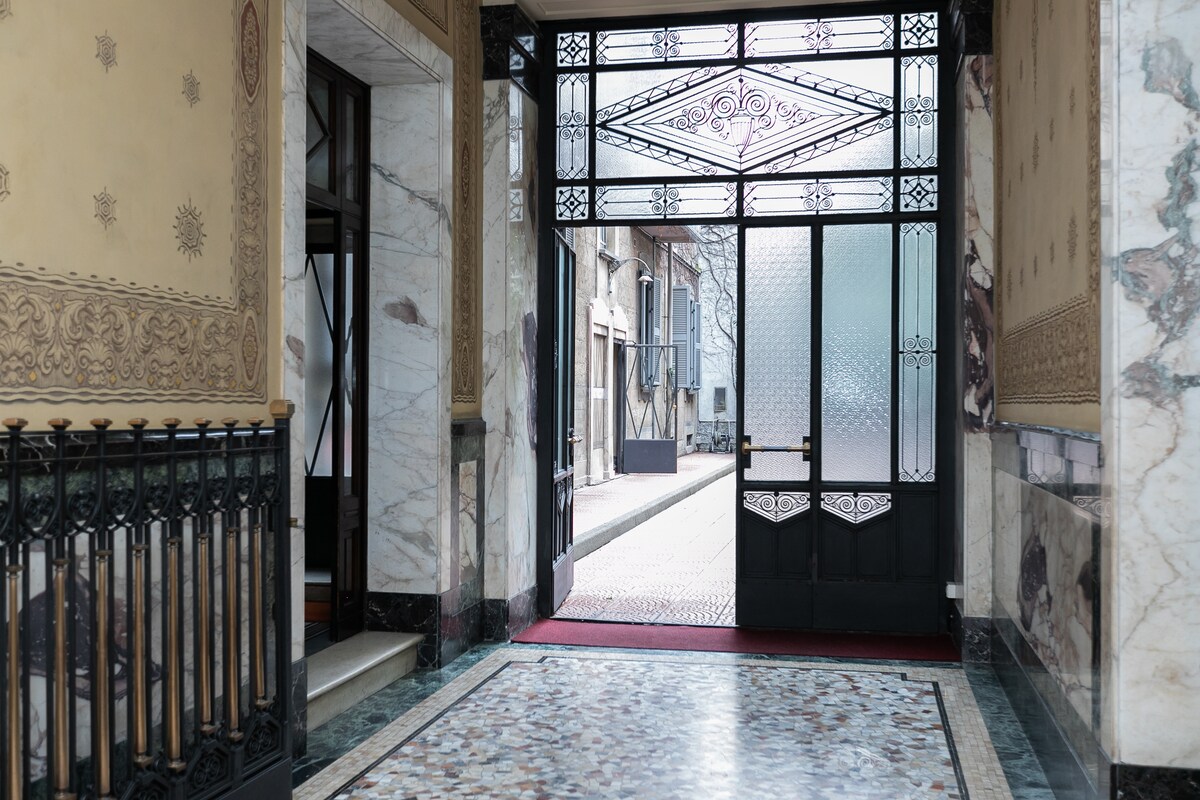 An elegant entryway features intricate marble flooring and decorative wall details. Tall doors framed with glass allow natural light to enter, highlighting the ornate architectural elements. A staircase is visible on the left, adding to the sophisticated ambiance of the space.