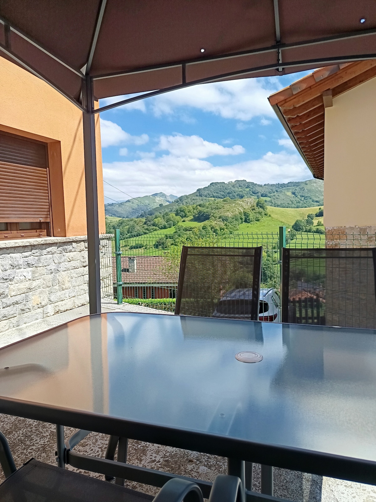 A covered terrace features a glass table surrounded by black chairs. The view reveals rolling green hills and mountains under a partly cloudy sky, offering a tranquil outdoor setting. A stone wall and neighboring buildings frame the area.