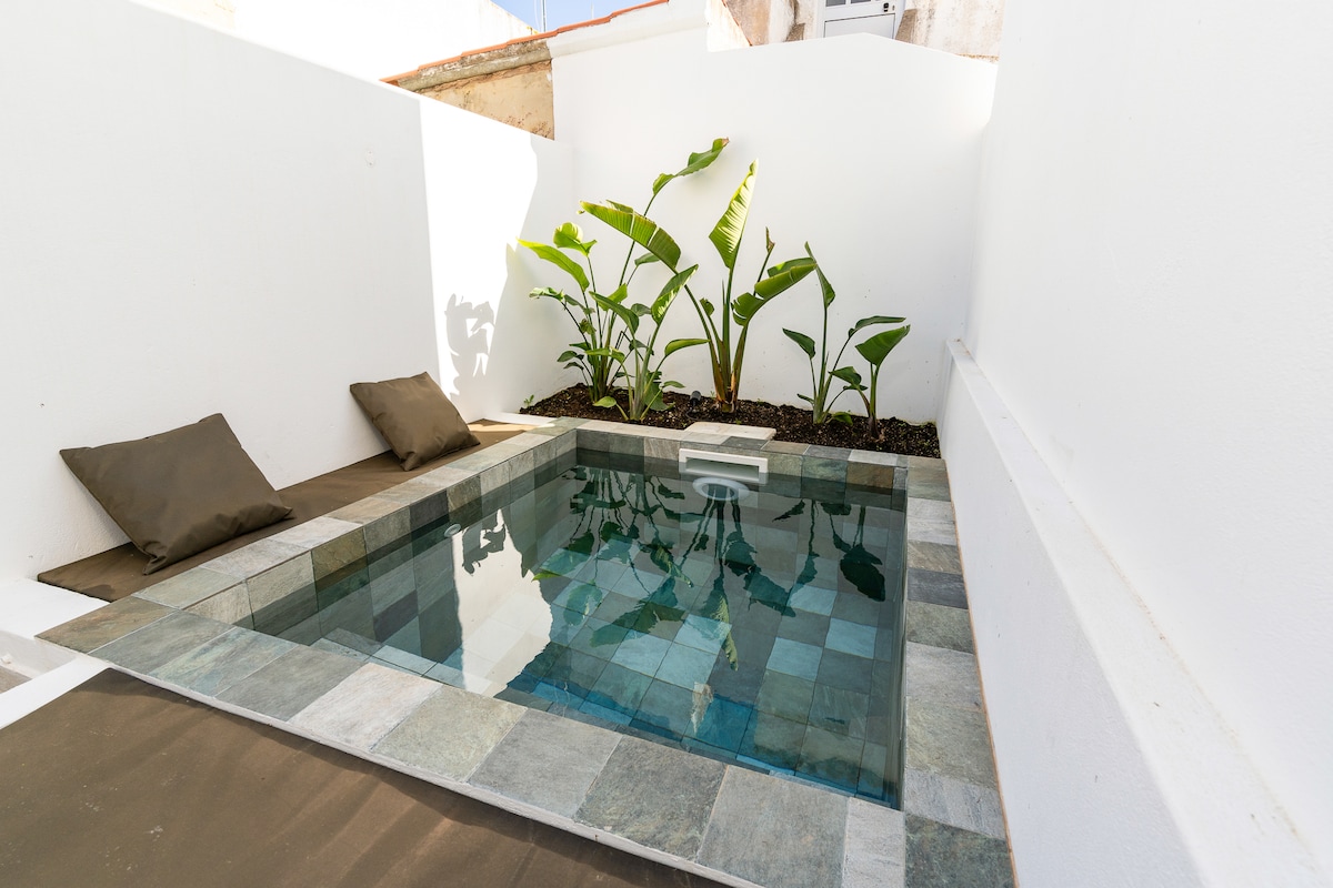 An intimate outdoor space features a small water pool surrounded by smooth stone tiles. Plush, neutral-colored cushions are placed on the side, and tropical plants add a touch of greenery against the white walls.