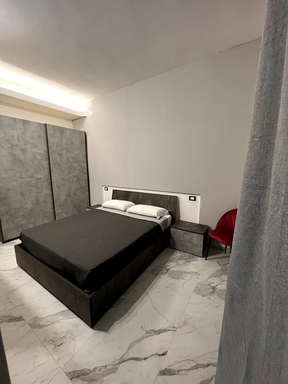 A modern bedroom features a spacious bed with a black coverlet, complemented by a simple bedside table. Smooth marble flooring reflects natural light. A subtle red chair is positioned beside the bed, and sliding closet doors provide a clean and uncluttered look to the space.