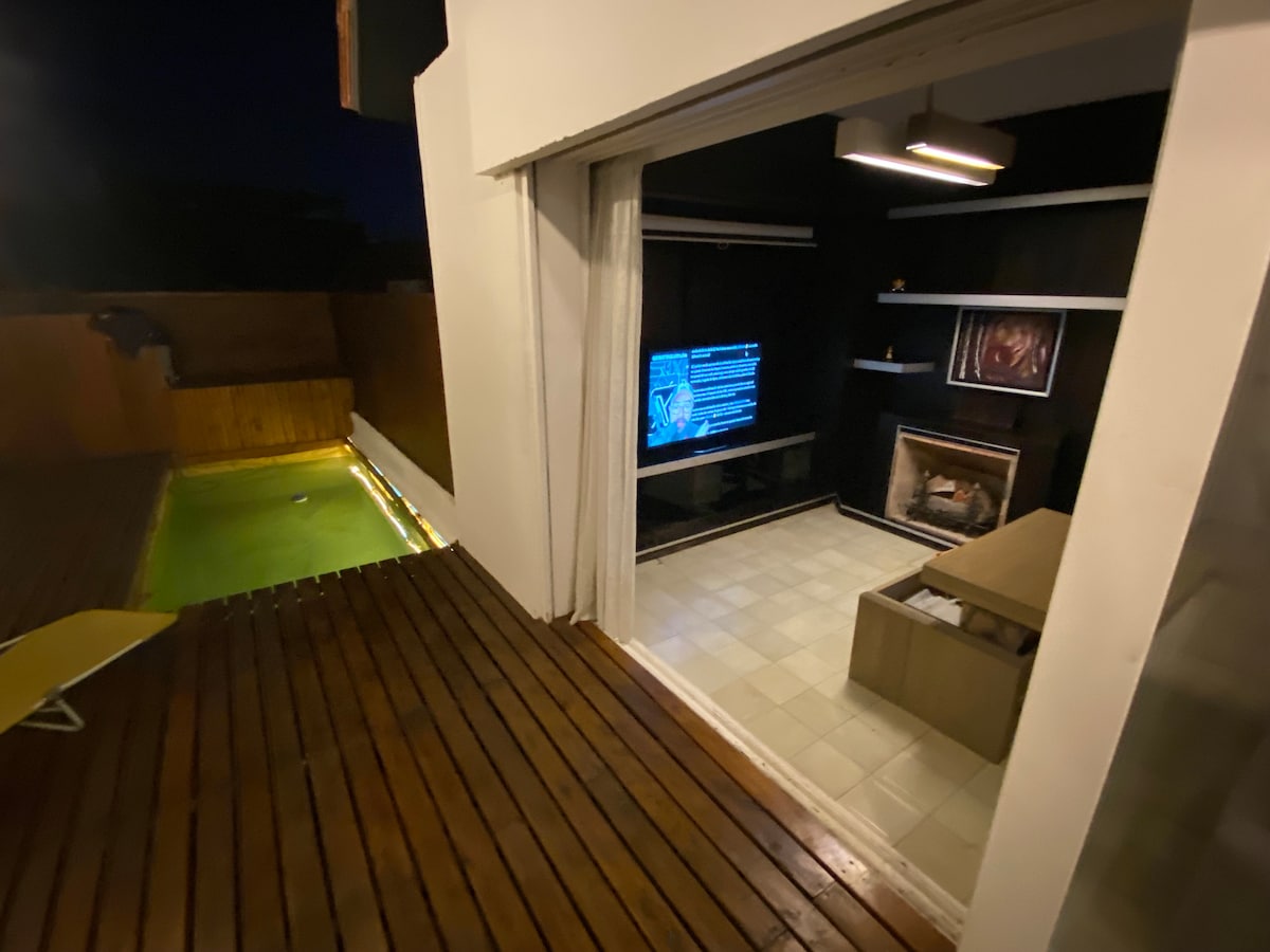 An indoor space features a television mounted on a dark wall and a cozy seating area with a coffee table. Outside the large sliding door, a pool is illuminated and a wooden deck extends alongside it, creating a seamless transition between indoor and outdoor areas.