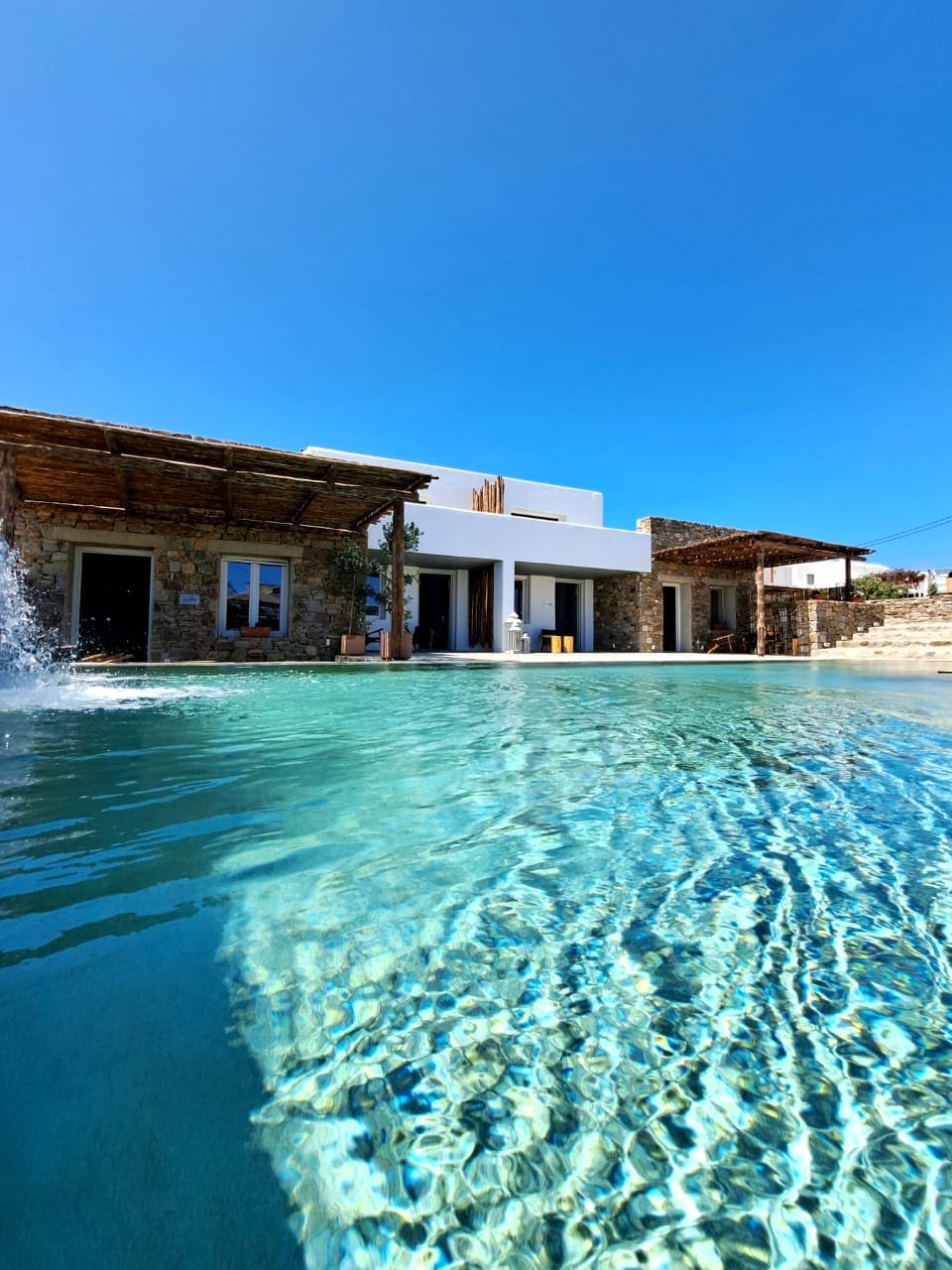 The image presents a poolside view featuring a crystal clear pool with gentle ripples. Surrounding the pool, stone and white-walled structures reflect traditional architecture, under a bright blue sky. Outdoor seating areas are visible beside the water, creating a serene atmosphere.