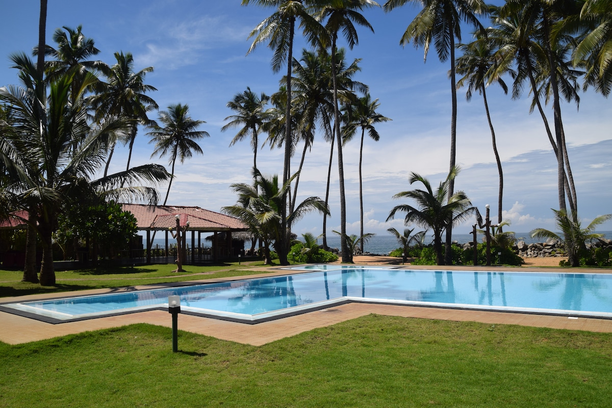 A serene outdoor area showcases a large swimming pool surrounded by lush green grass and palm trees. The pool reflects the blue sky above, while a charming gazebo can be seen nearby, offering shaded seating for relaxation.