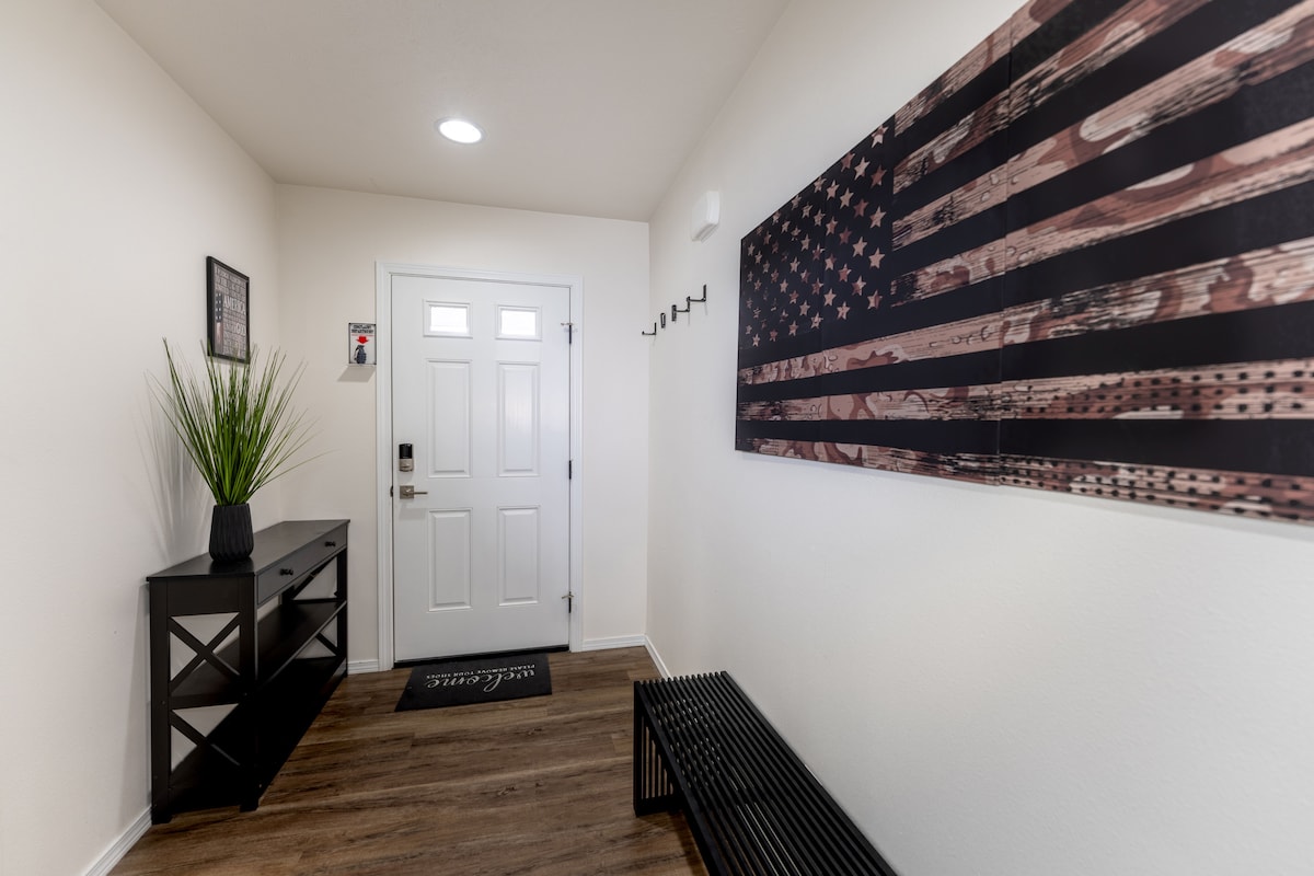 An inviting entryway features a light-colored wall and a stylish wooden floor. A console table sits against one wall, complemented by a decorative plant. A large wall art piece depicting an American flag adds character, while a coat rack provides space for hanging belongings.