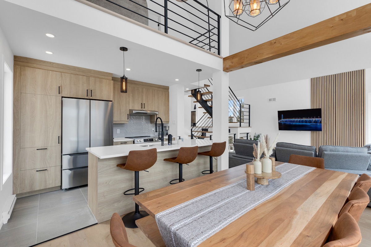 A modern kitchen and dining area are depicted, featuring a large wooden table surrounded by comfortable chairs. A sleek kitchen island with bar stools and stainless-steel appliances complements the wood cabinetry. Natural light fills the space, enhancing the airy atmosphere.