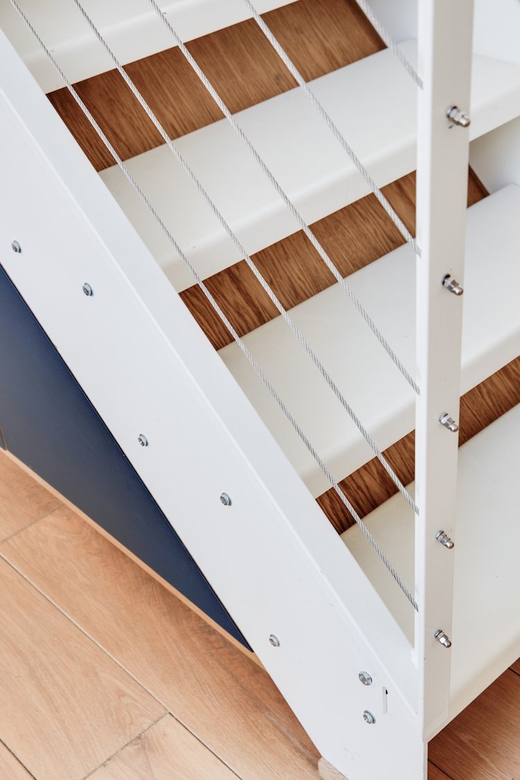 A modern staircase is depicted, featuring white steps and a sleek wooden accent. The railings are designed with metal cables, providing an open, airy feel. Light wood flooring extends across the area, enhancing the contemporary aesthetic.