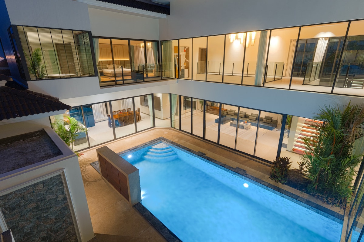 A view of the villa's private pool surrounded by modern architecture. Large glass doors provide visibility into the open living areas. The pool features gentle steps and is illuminated by soft lighting, complemented by tropical plants in the surrounding garden.