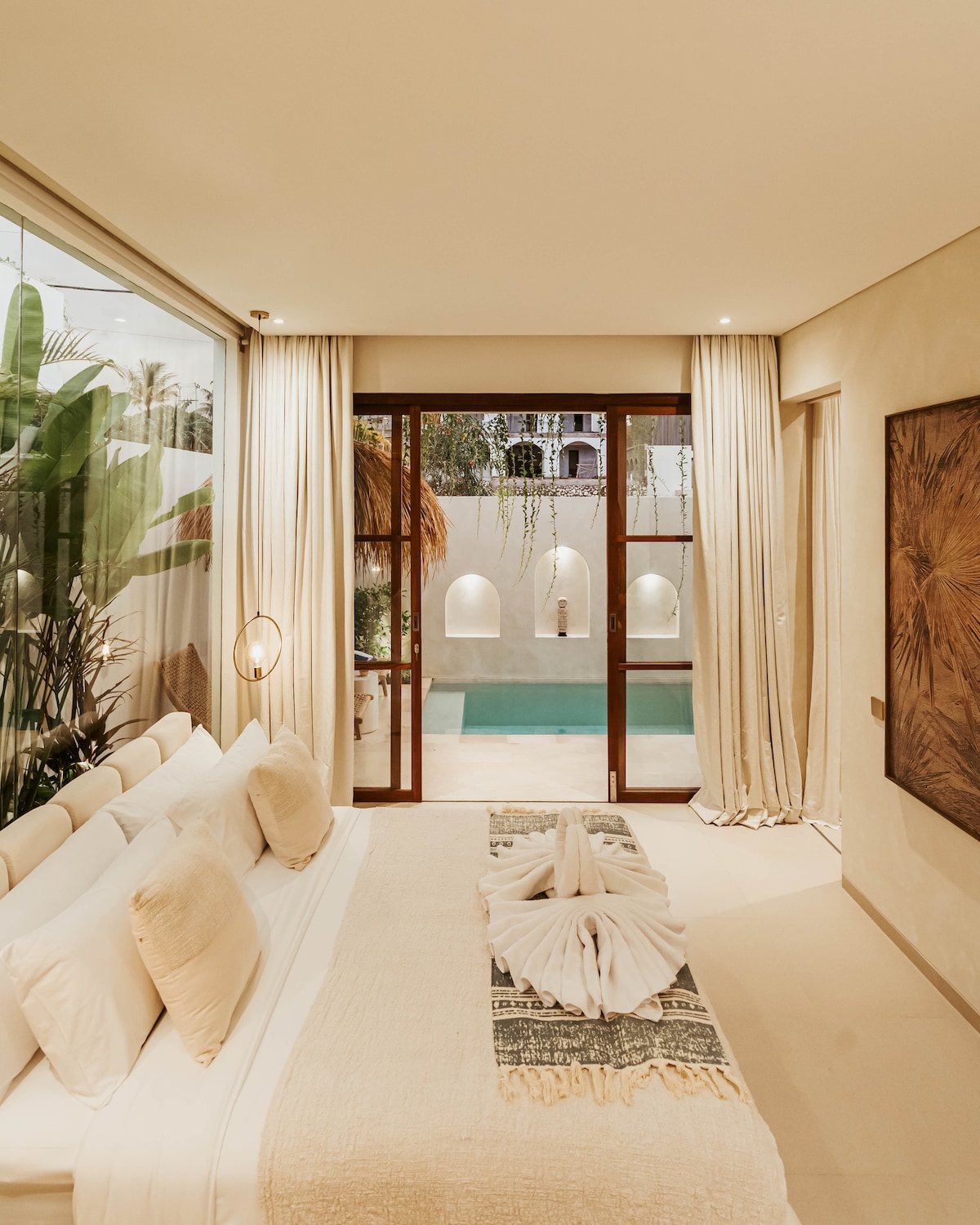 A serene bedroom features a spacious bed adorned with neutral-toned linens. Large glass doors lead to a private pool area, surrounded by tropical greenery and soft ambient lighting, creating a calm and inviting atmosphere.