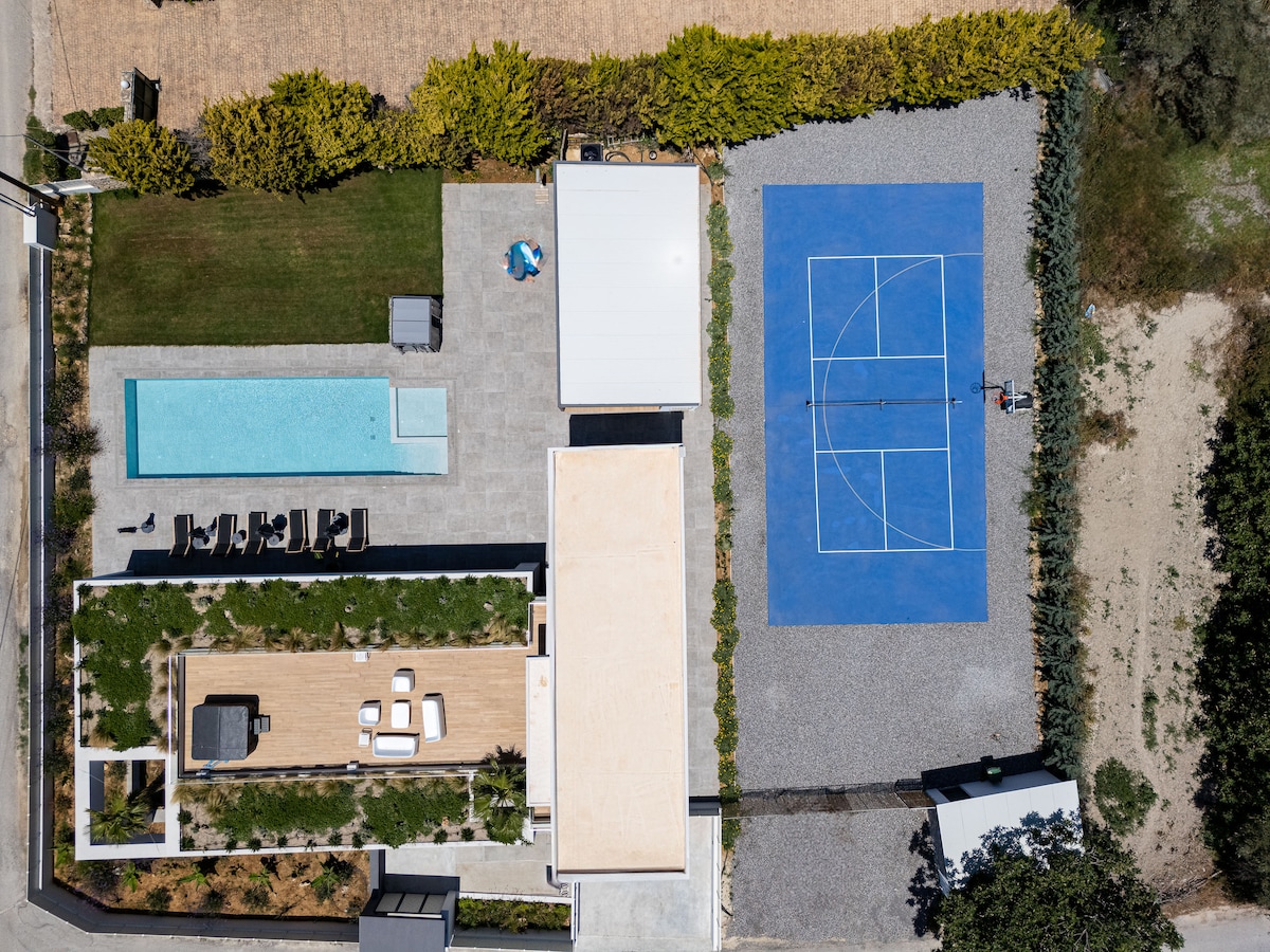 An aerial view showcases the outdoor amenities of Manza Villa, featuring a long saltwater pool adjacent to a spacious wooden deck with loungers. A brightly colored pickleball court is visible alongside the neatly landscaped yard, highlighting the villa's recreational options.
