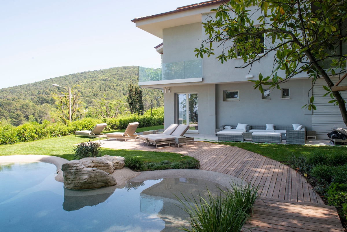 An outdoor area features a serene pool with natural stones and wooden decking. Comfortable lounge chairs are positioned beside the pool, while a shaded seating arrangement provides additional relaxation space. Lush greenery and gentle hills create a tranquil backdrop beyond the property.