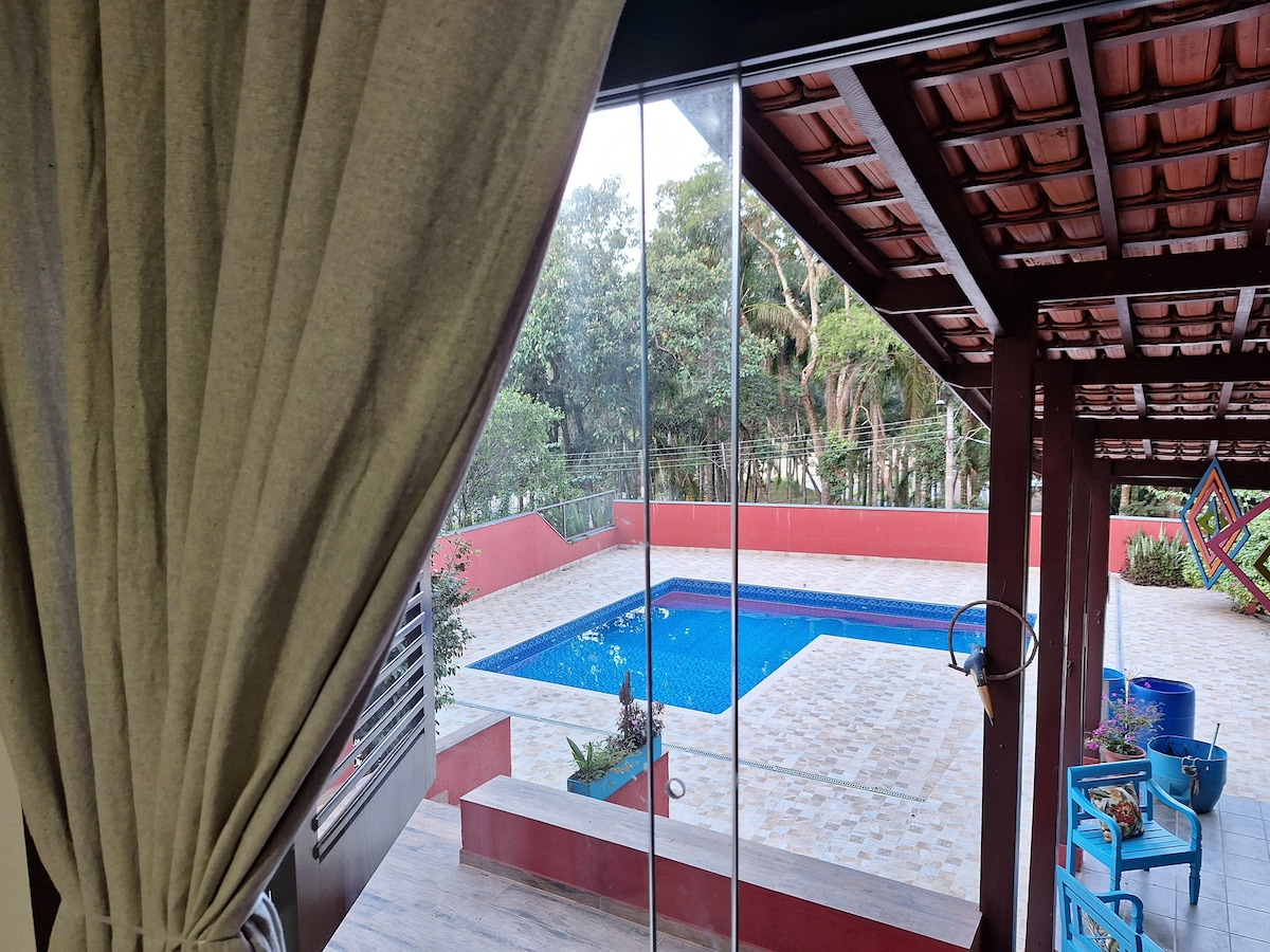 An inviting view of a swimming pool is visible through a large glass window. The pool area is surrounded by vibrant landscaping and features a spacious tiled deck. Soft curtains frame the view, enhancing the peaceful atmosphere of the outdoor space.