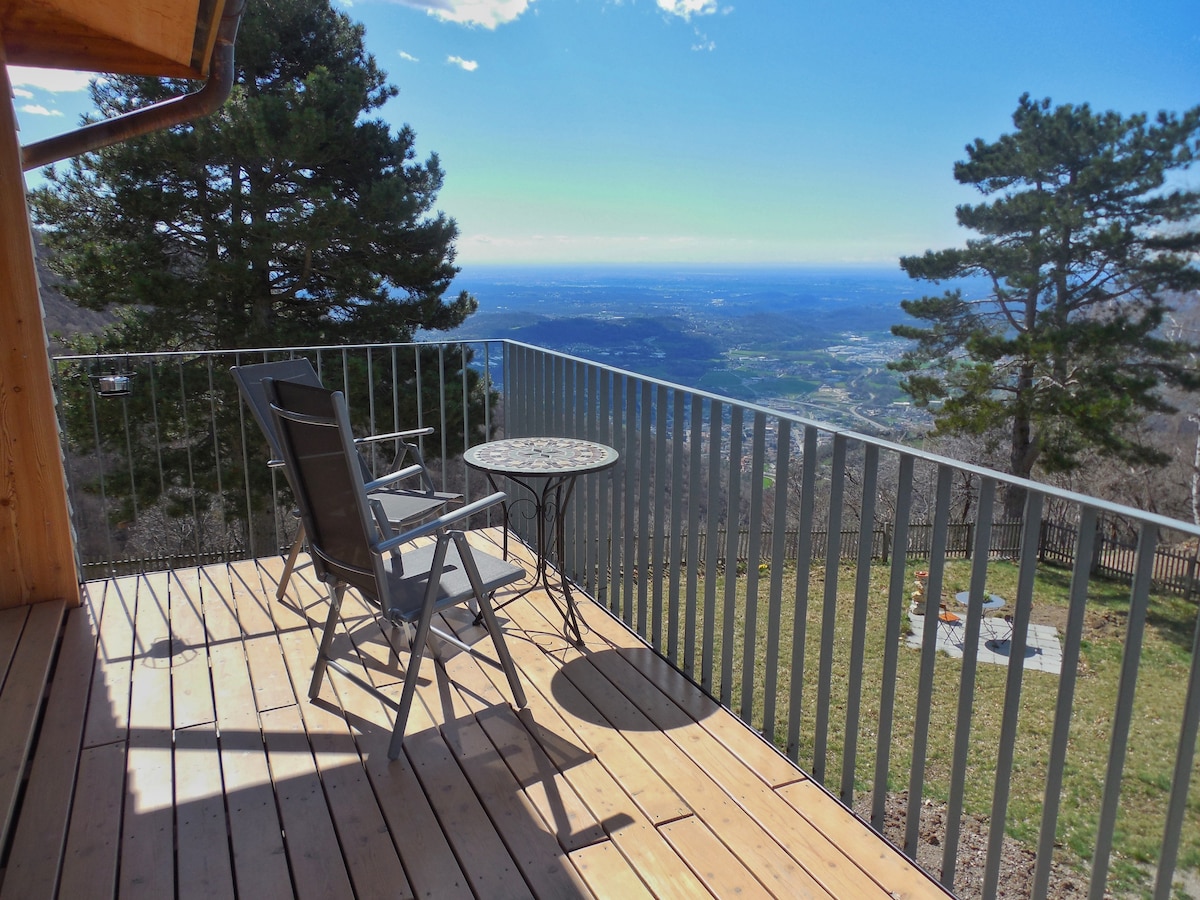 A wooden terrace is equipped with a small round table and a lounge chair, offering panoramic views of the valley below. Tall pine trees frame the space, and the clear sky enhances the natural setting.