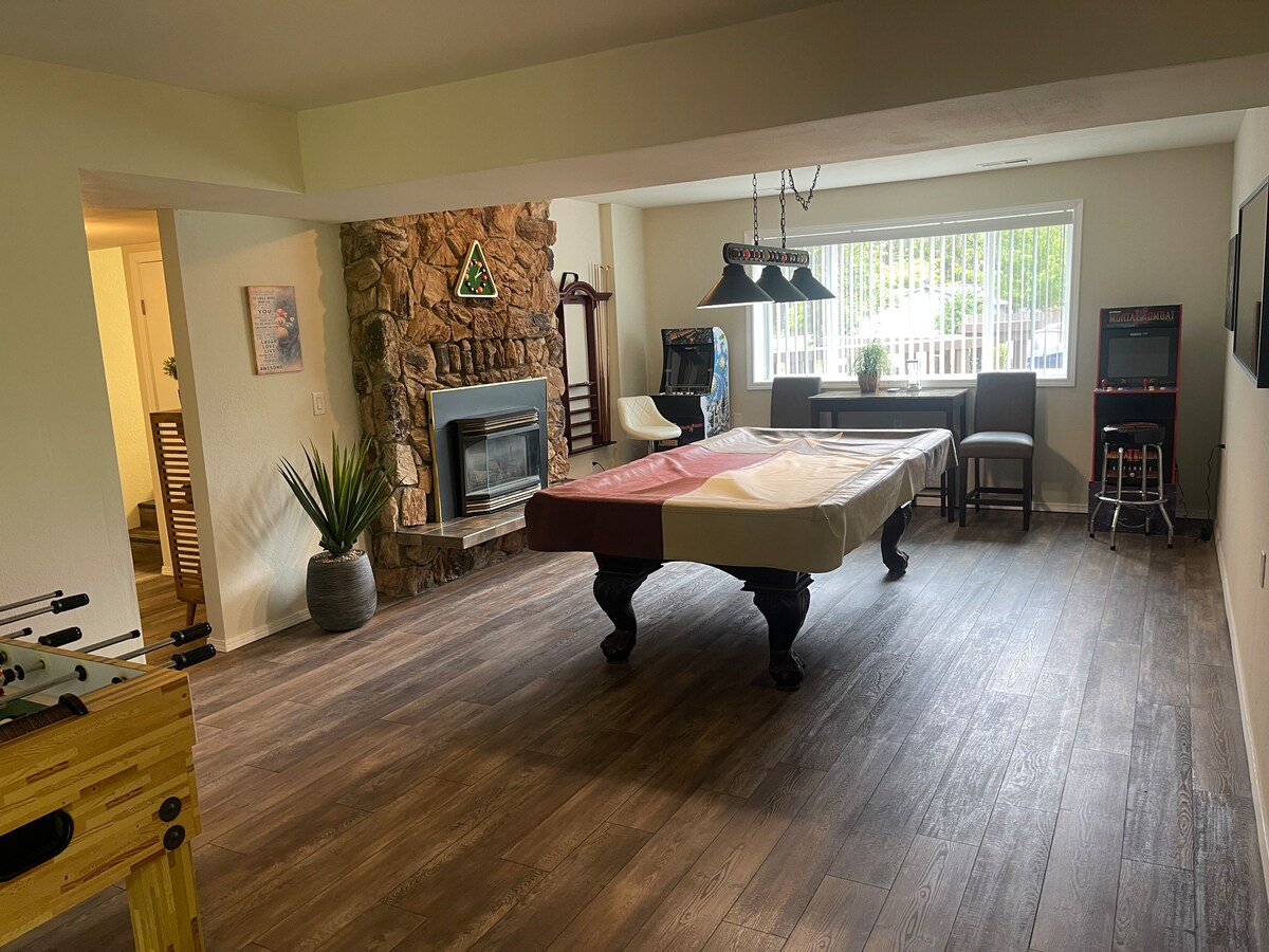 A spacious game room is depicted with a pool table covered partially, alongside a foosball table. A stone fireplace and large window provide illumination, while arcade games are visible in the corner. Comfortable seating is arranged nearby, contributing to a relaxed environment.