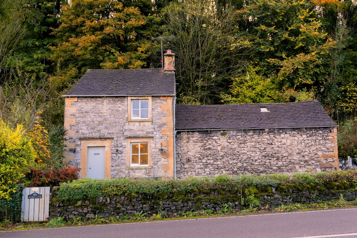 The exterior of a charming stone cottage is visible, accented by a slate roof and lush greenery surrounding it. A low stone wall lines the property, with vibrant autumn foliage providing a seasonal touch. The entrance features a simple door framed by a small window.