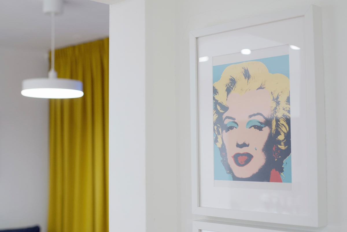 A framed artwork is displayed on a white wall, with soft colors and abstract design partially visible. A bright pendant light hangs above, illuminating the space. Behind, flowing yellow curtains add a touch of vibrancy, creating a welcoming environment.