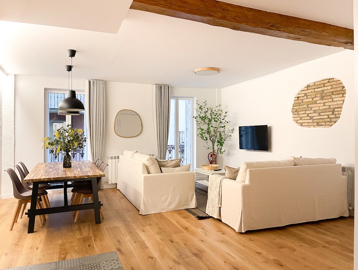 A spacious living and dining area features neutral-toned sofas and a large wooden dining table surrounded by chairs. Natural light enters through balcony doors, illuminating the space. Decorative plants add greenery, while a flat-screen TV is mounted on the wall. Warm wood accents enhance the atmosphere.