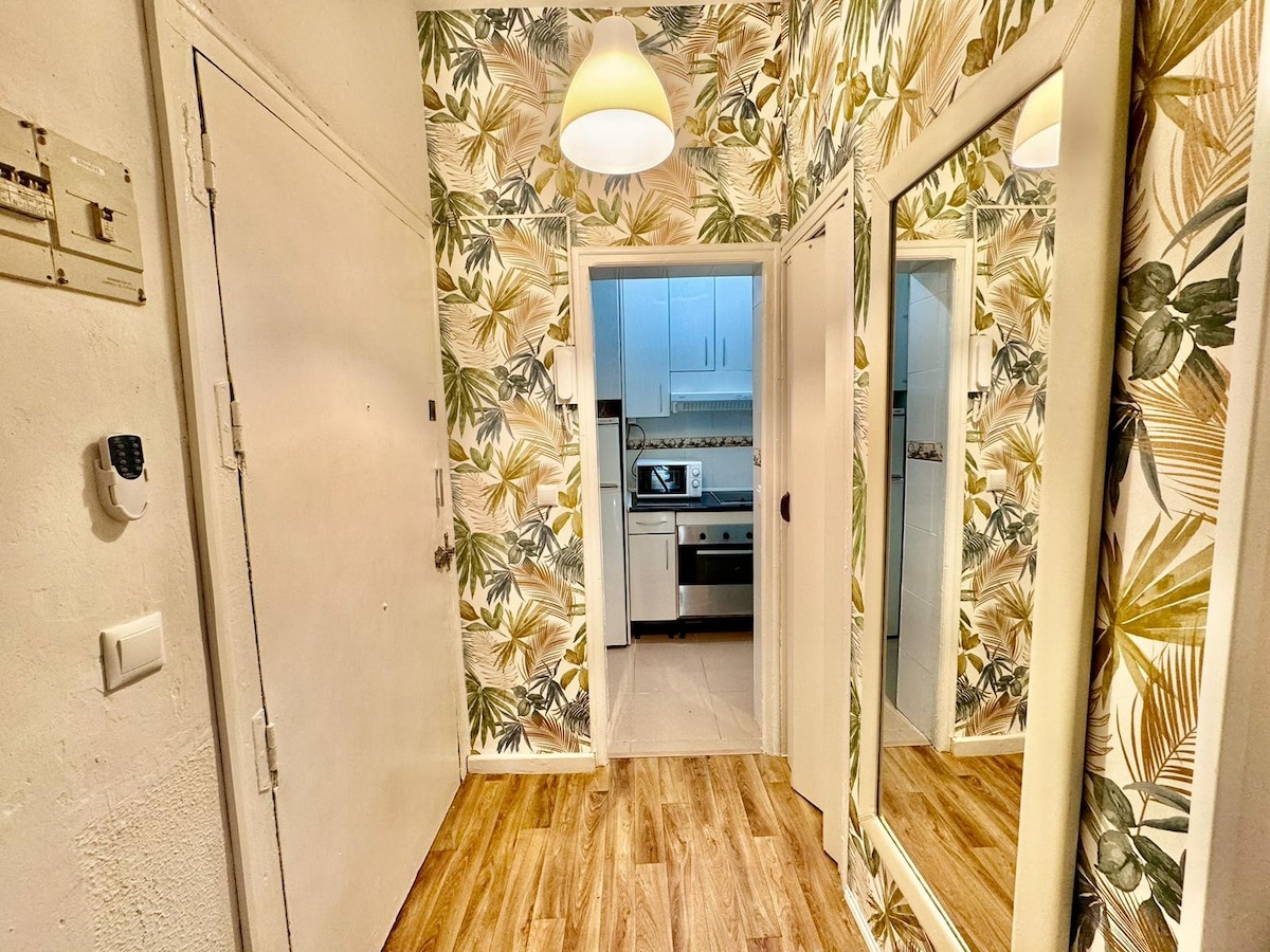 An entryway is highlighted with tropical-themed wallpaper, featuring leafy designs in green and gold. A light fixture hangs from the ceiling, casting a soft glow. Two mirrors create an open feel while doors lead to a kitchen area visible in the background.
