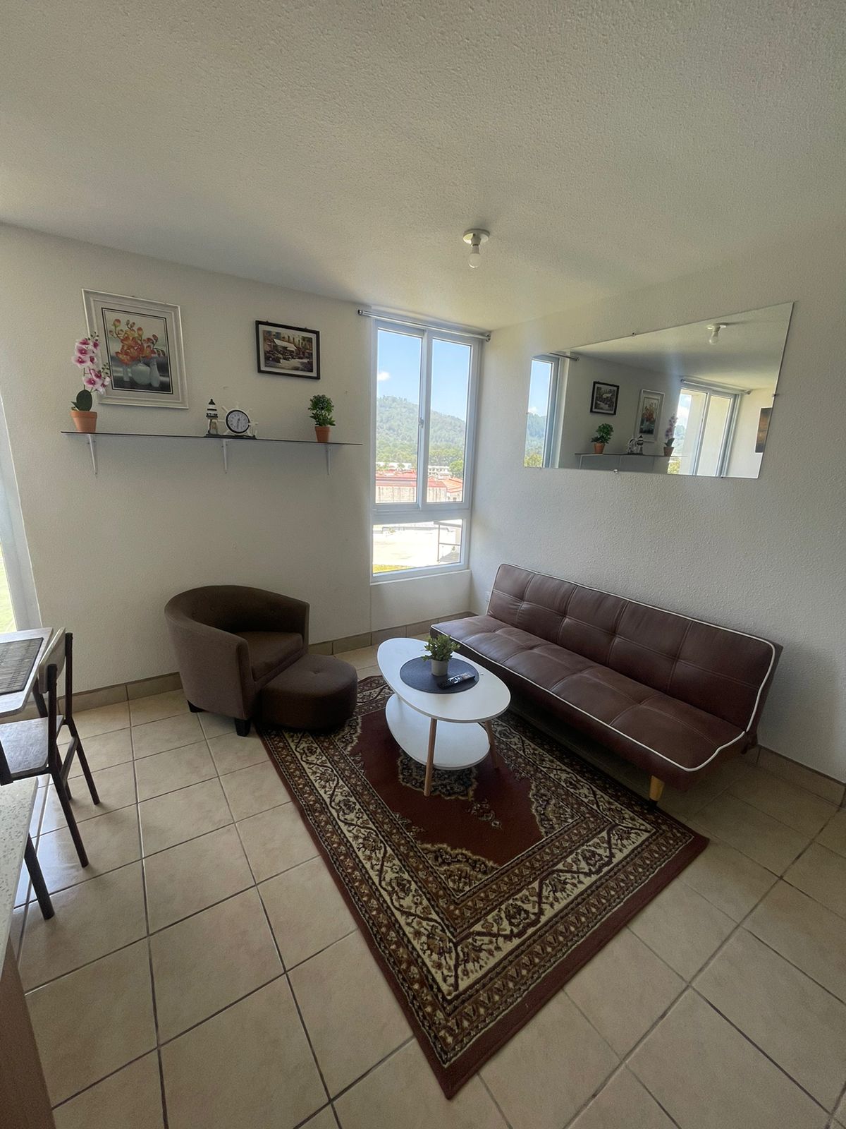 A cozy living area is presented, featuring a brown futon and a round white table. A single brown armchair complements the space. Light enters through large windows, casting a bright ambiance. Decorative plants and artwork enhance the walls, contributing to a welcoming atmosphere.