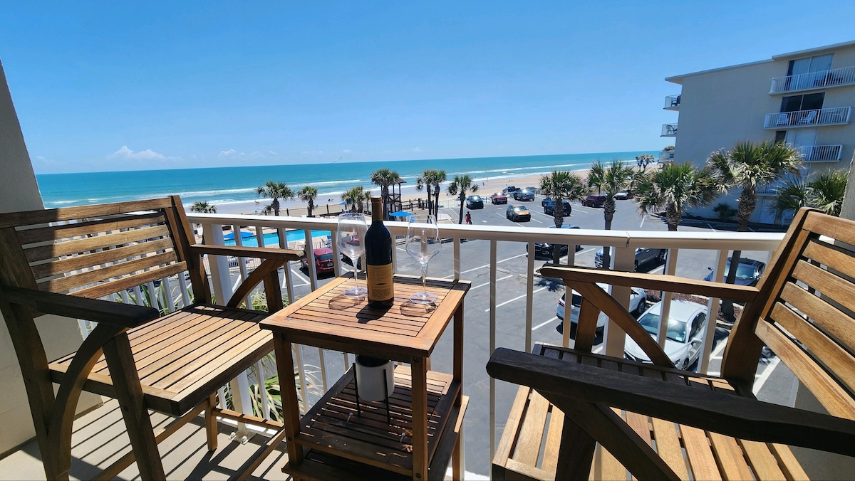 A balcony seating area is featured, with two wooden chairs and a small table set. A bottle of wine and glasses are placed on the table. The view overlooks the ocean and sandy beach beyond, with palm trees lining the area and parked cars visible below.