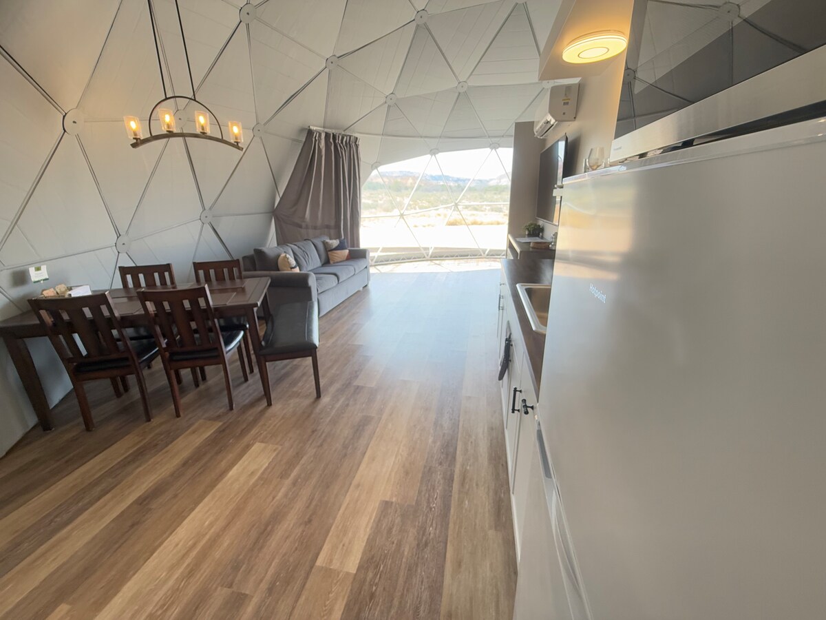 An open living area is presented with a spacious sitting area featuring a grey sofa and a dining table surrounded by wooden chairs. Natural light enters through large windows, offering views of the landscape. A kitchen area is equipped with modern appliances.