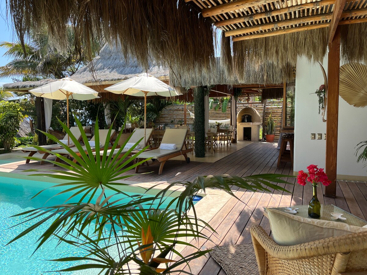 A tranquil outdoor area features a shimmering pool surrounded by loungers and shaded by umbrellas. An inviting dining space is visible in the background, with a rustic outdoor grill nearby. Tropical plants add a touch of greenery, harmonizing with the natural decor.