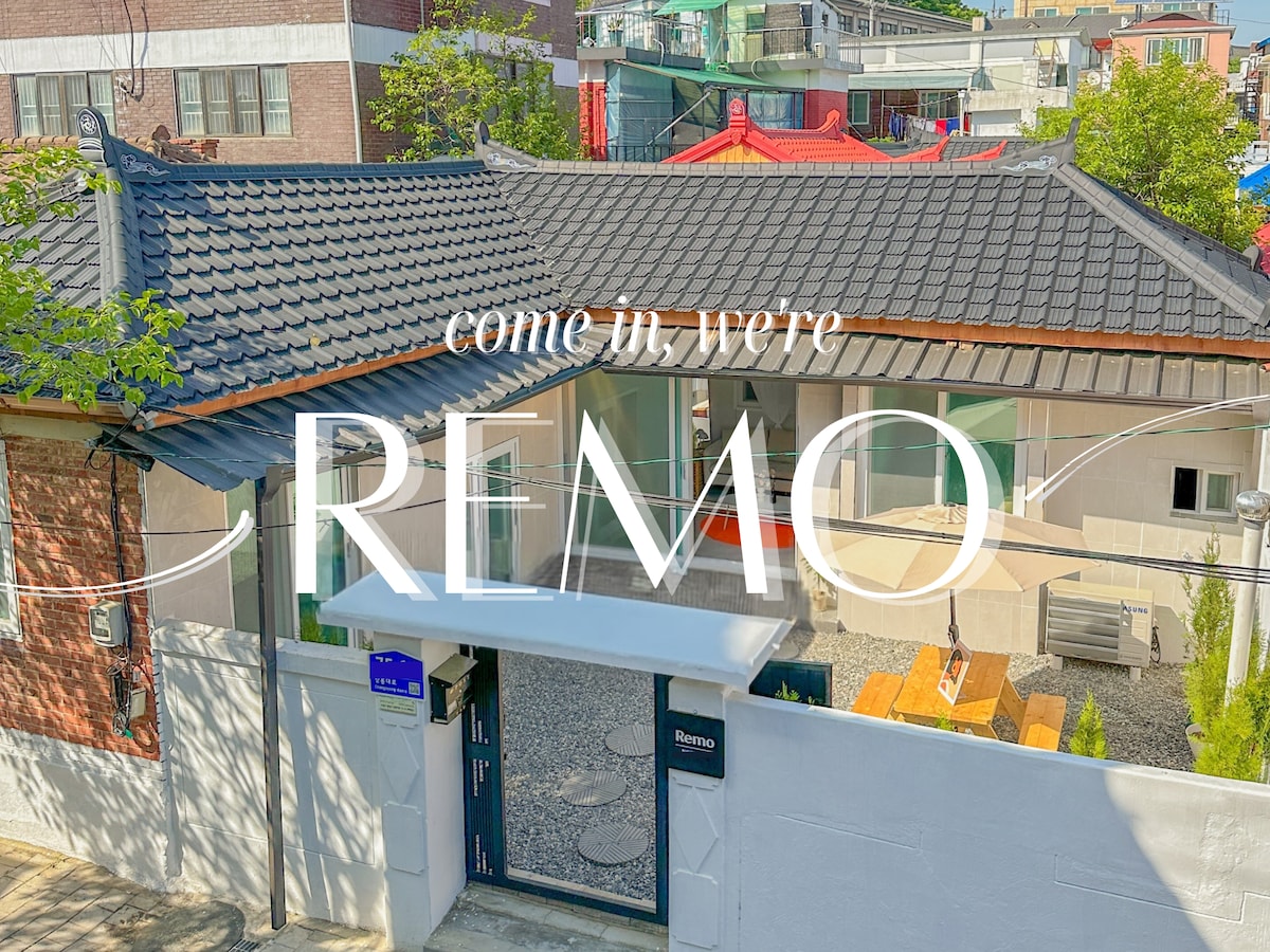 The exterior of a remodeled hanok showcases a traditional tiled roof complemented by modern design elements. A welcoming entrance is visible with a clear view of a spacious courtyard surrounded by greenery, inviting guests to experience the unique charm of this accommodation.