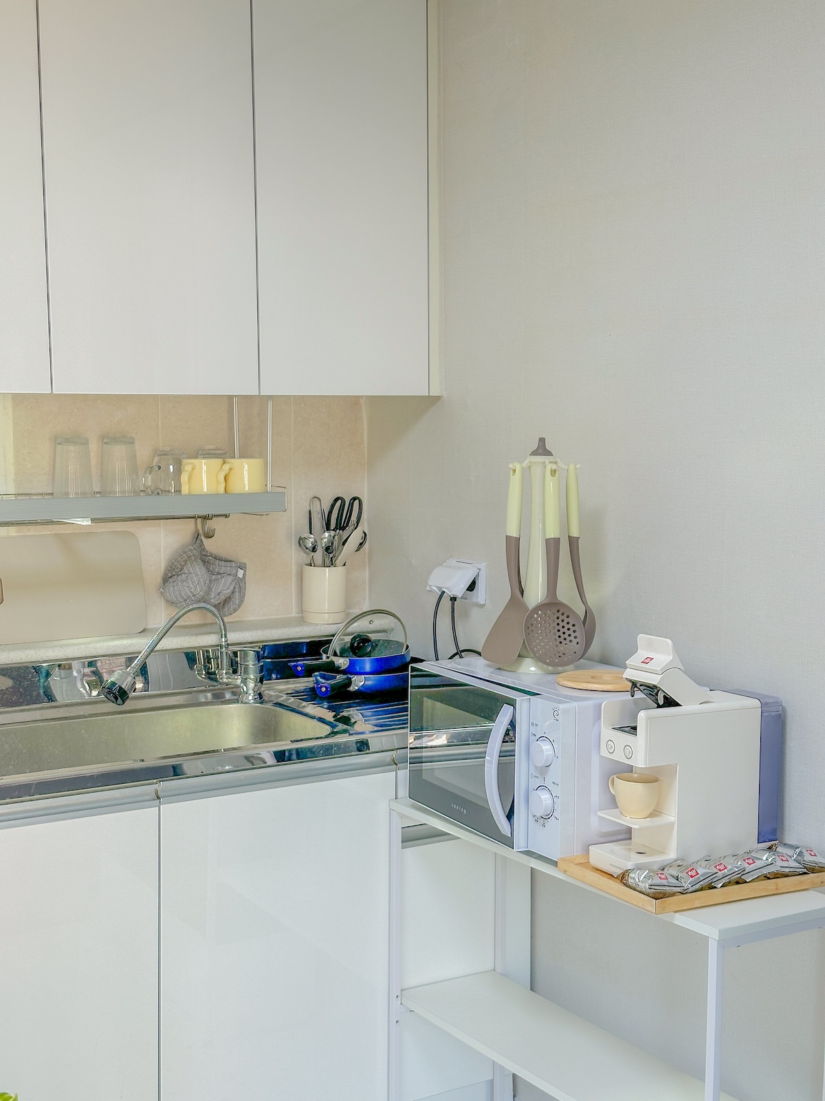A modern kitchen area is equipped with sleek cabinetry, featuring a microwave and coffee maker on a light wooden shelf. Stainless steel utensils are neatly organized, and a blue kettle sits on the stovetop. Transparent jars are placed on a surrounding countertop, enhancing functionality.