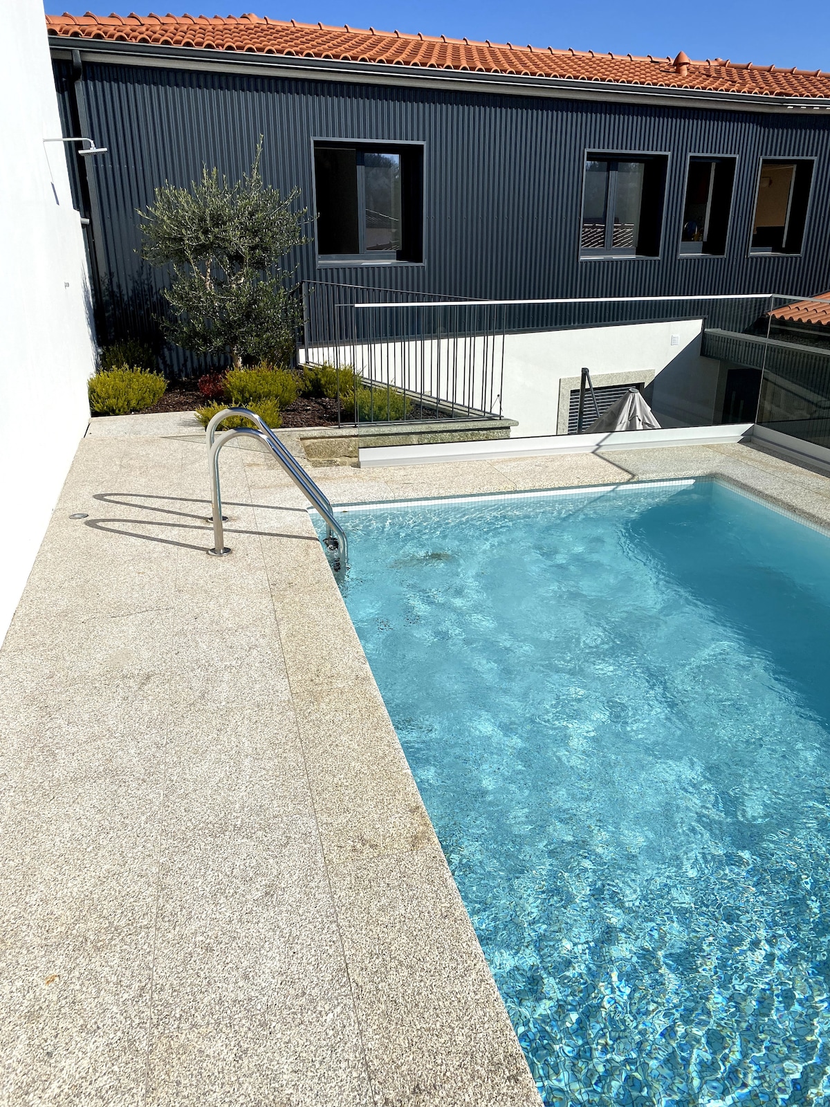 A heated pool is situated adjacent to a modern building, featuring a clear blue surface. Smooth gray stone surrounds the pool, with a metal railing providing safety. Lush greenery is visible at the pool’s edge, contributing to a serene outdoor space.